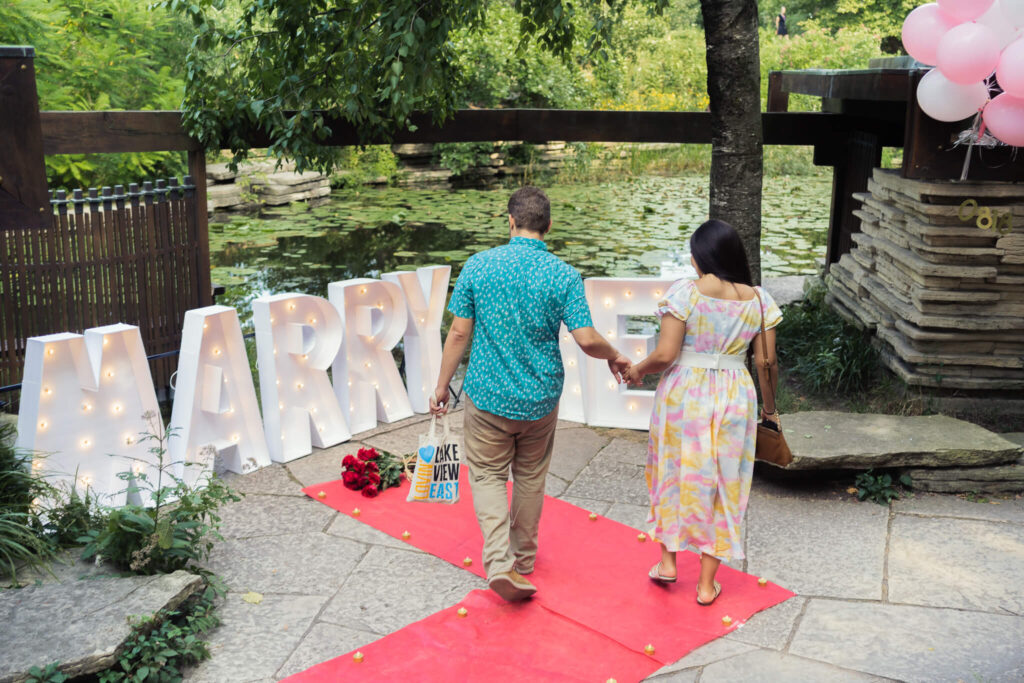 Holding hands, a couple approaches large "Marry Me" letters perched on the edge of the Lily Pool. There is a red carpet with flowers at their feet.