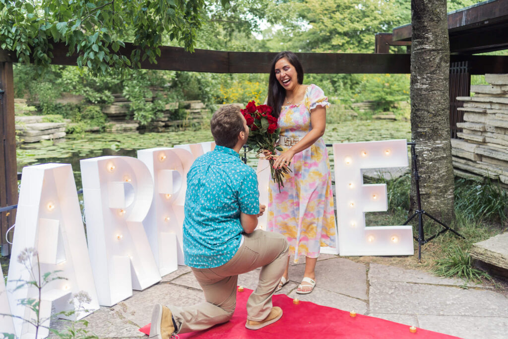 A proposal in action! A man in a blue shirt is down on one knee in front of a woman with a mid-length colorful dress. She is delighted, her mouth open in ecstasy. 