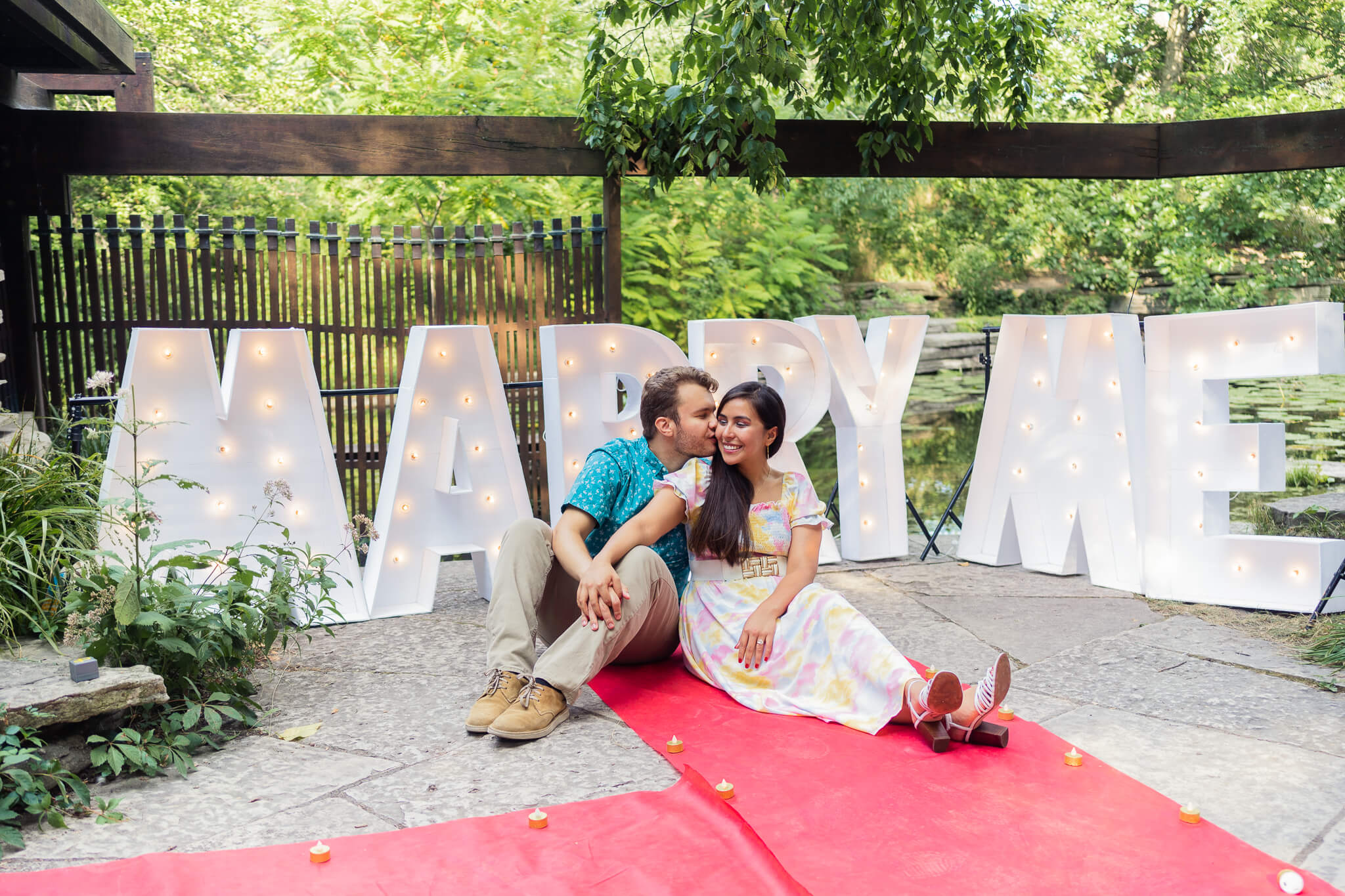 A couple sits in front of large "marry me" letters. They are on a red carpet.