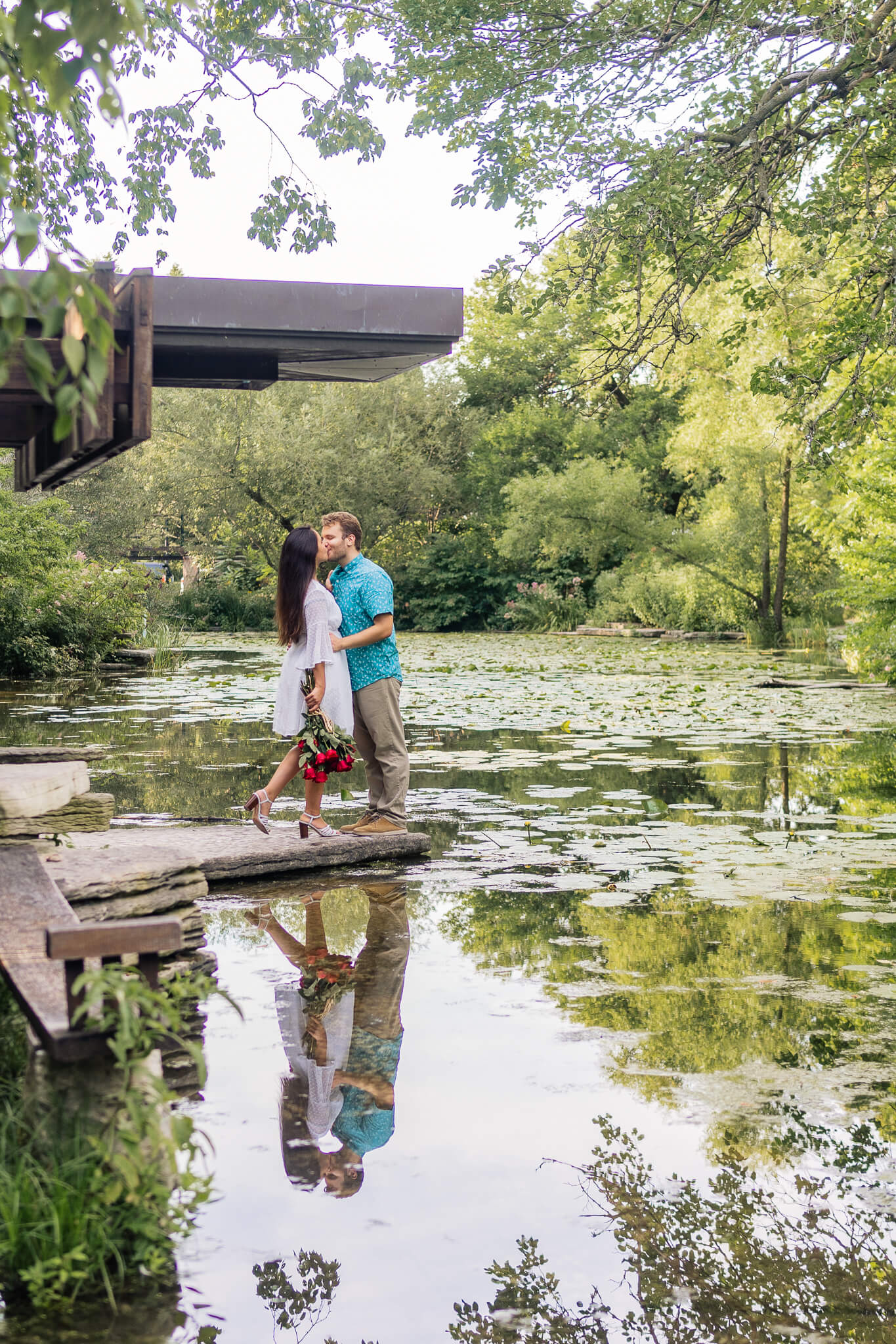 A woman kisses a man and pops her foot up. The couple is standing in the middle of the pond on a rock.