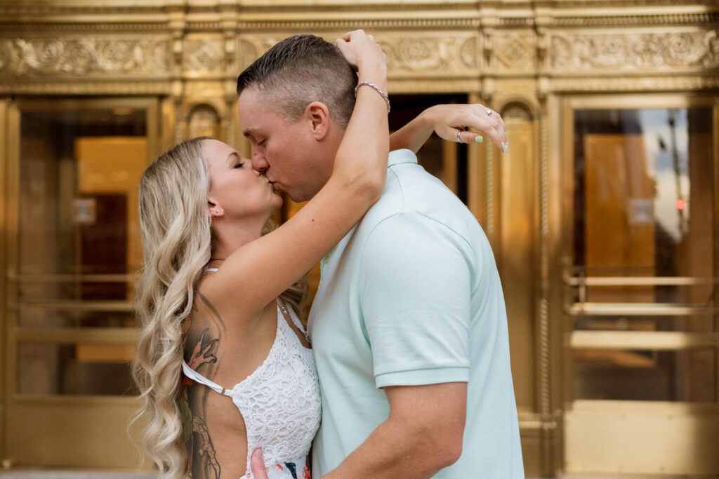A couple kisses in front of the wrigley building. Her hands are draped around his neck and her back tattoos are visible.