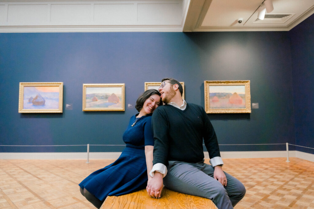 A man and woman sit back to back in a gallery at the art institutee of chicago, they lean back and smile at each other