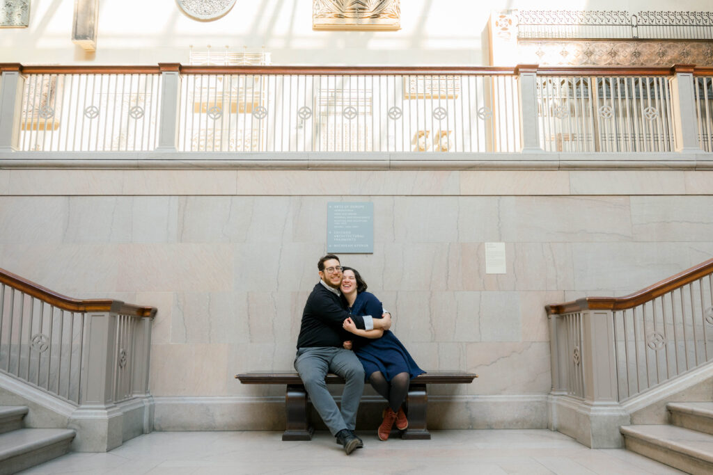 A man and woman smile and hug on a bench at their engagement session at the Art Institute of Chicago