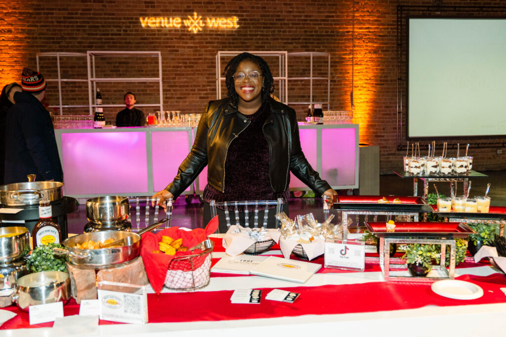 A woman representing Sweet Baby Ray's Catering smiles behind a table of food 