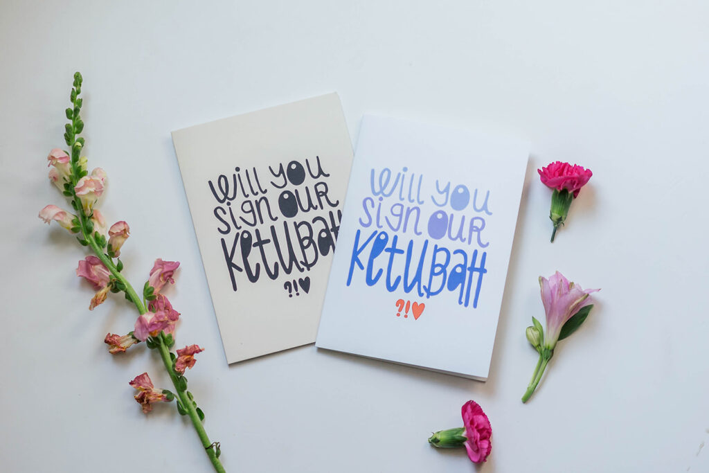 Two greeting cards each read "Will you sign our ketubah?" Three pink flowers lie below the cards.