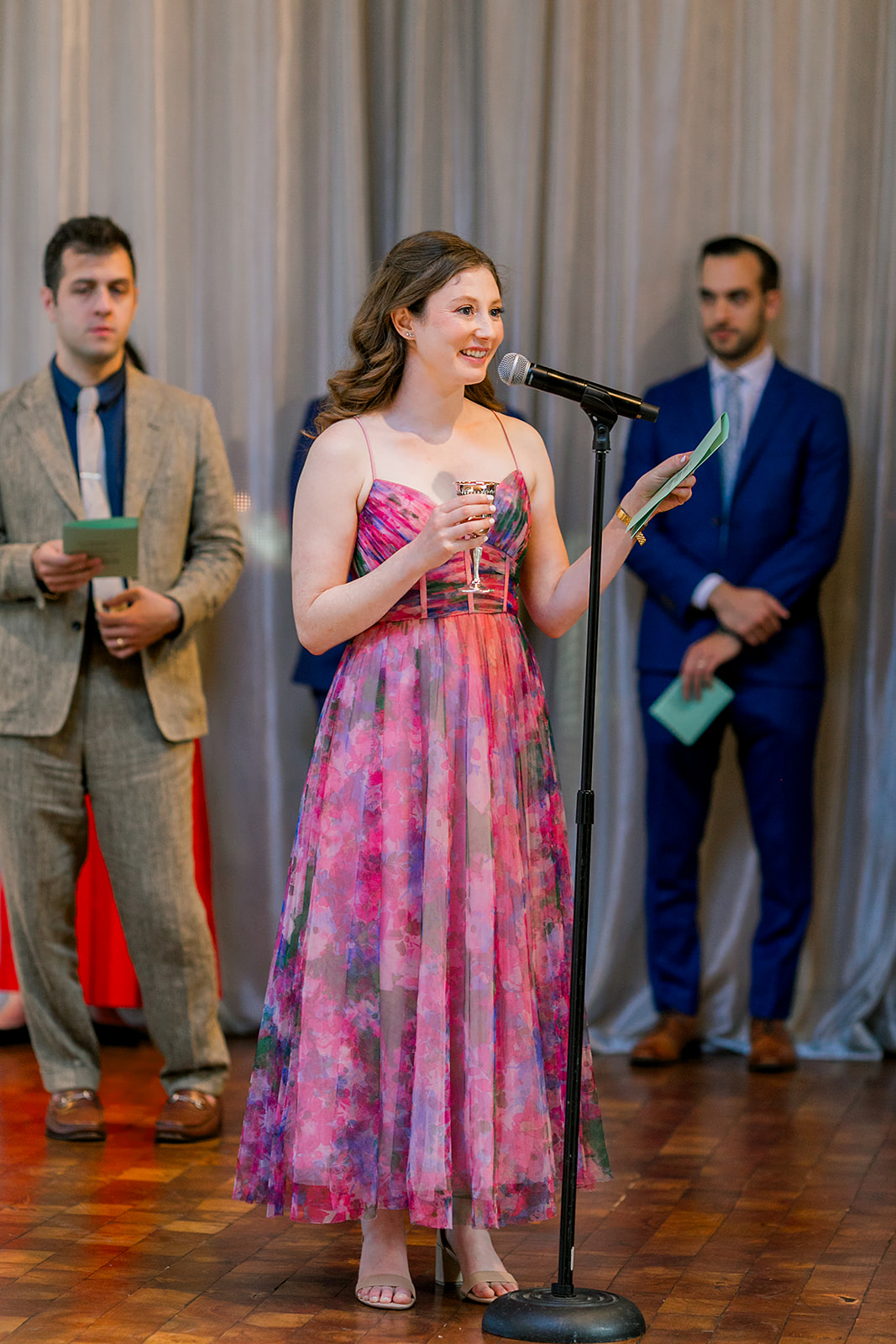 A woman in a pink dress gives a speech into the microphone at a Jewish wedding reception