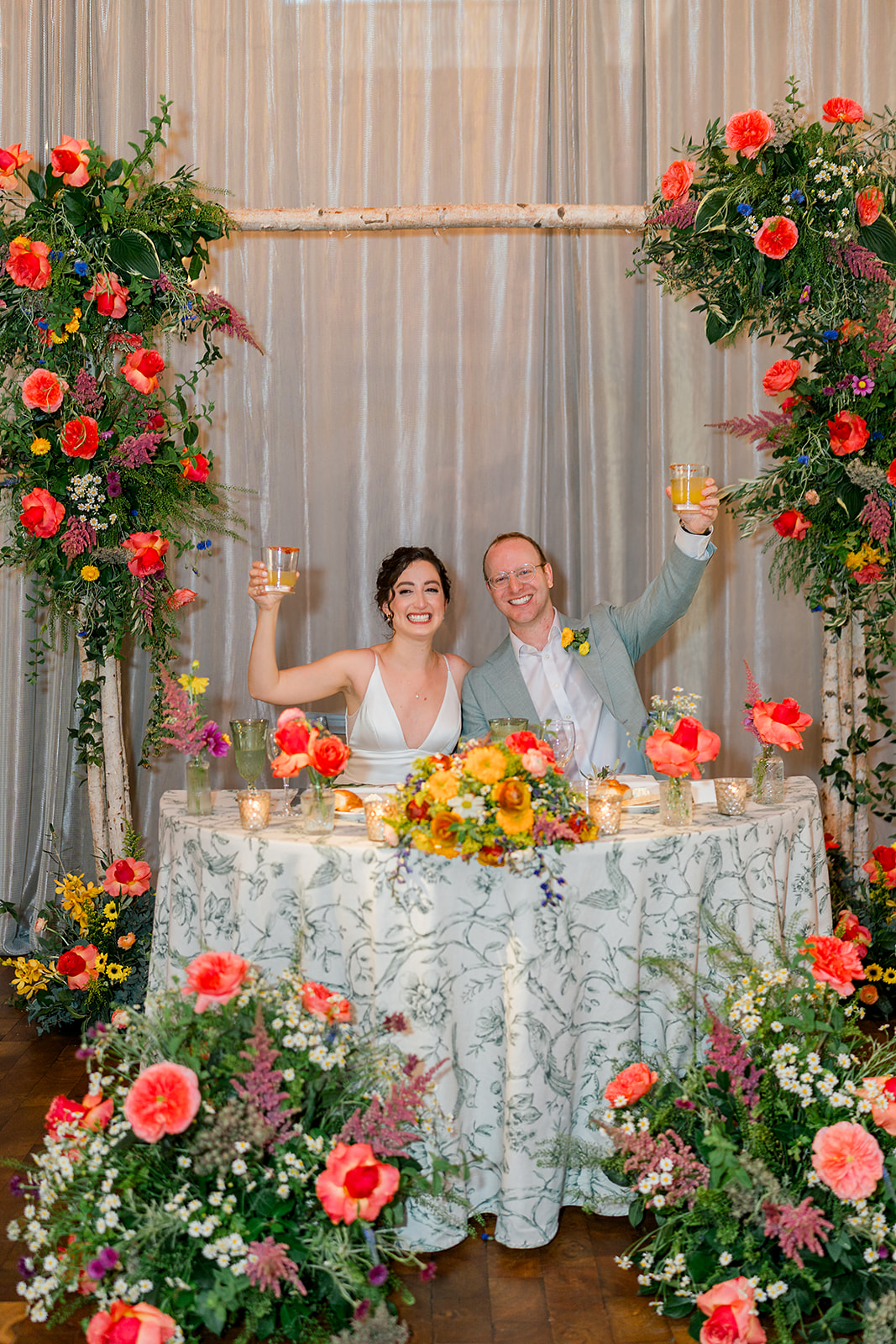 Newlyweds raise a glass at their vibrant floral covered head table during their Jewish wedding reception