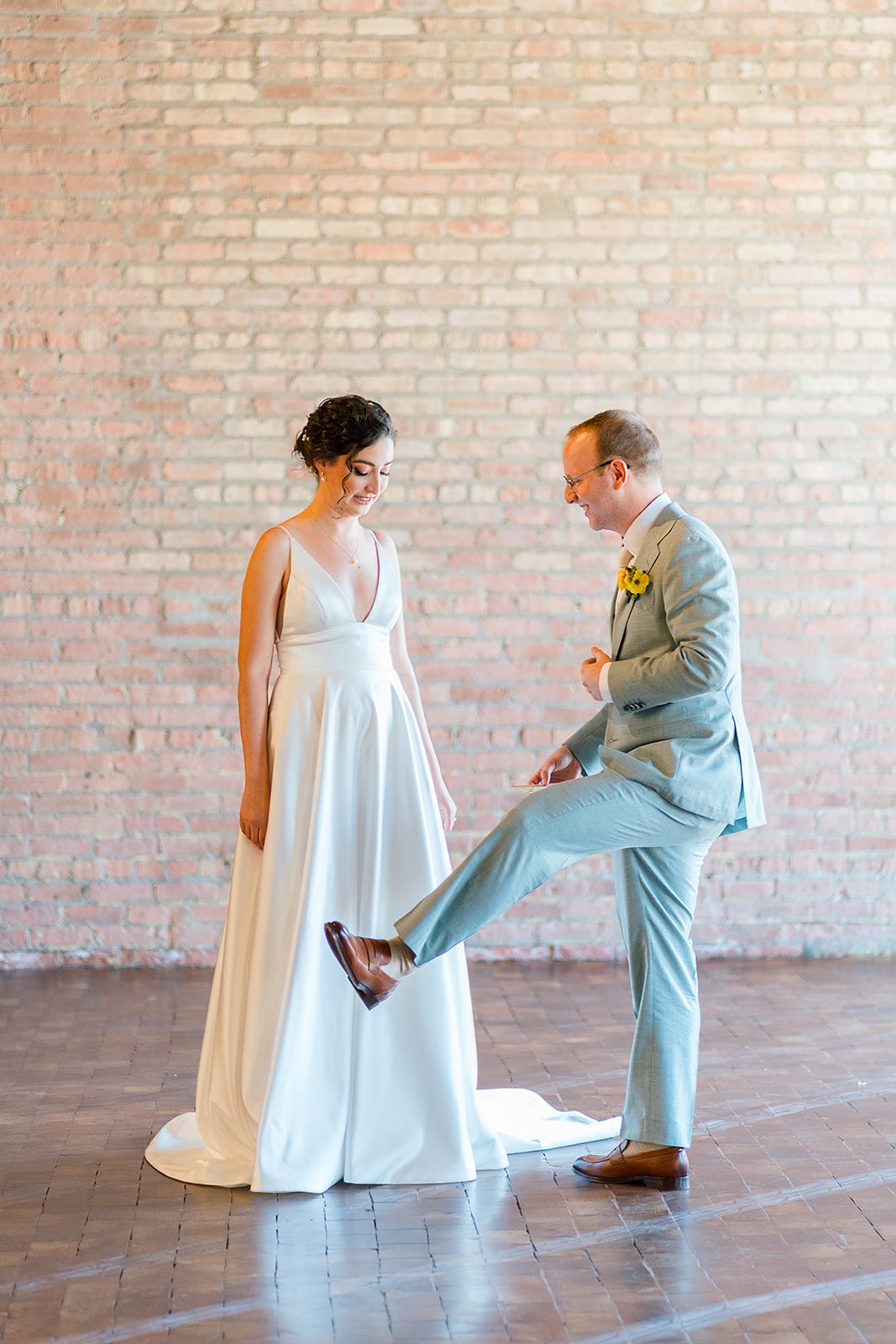 A groom in a green suit shows off his brown leather shoes to his bride during their Jewish wedding