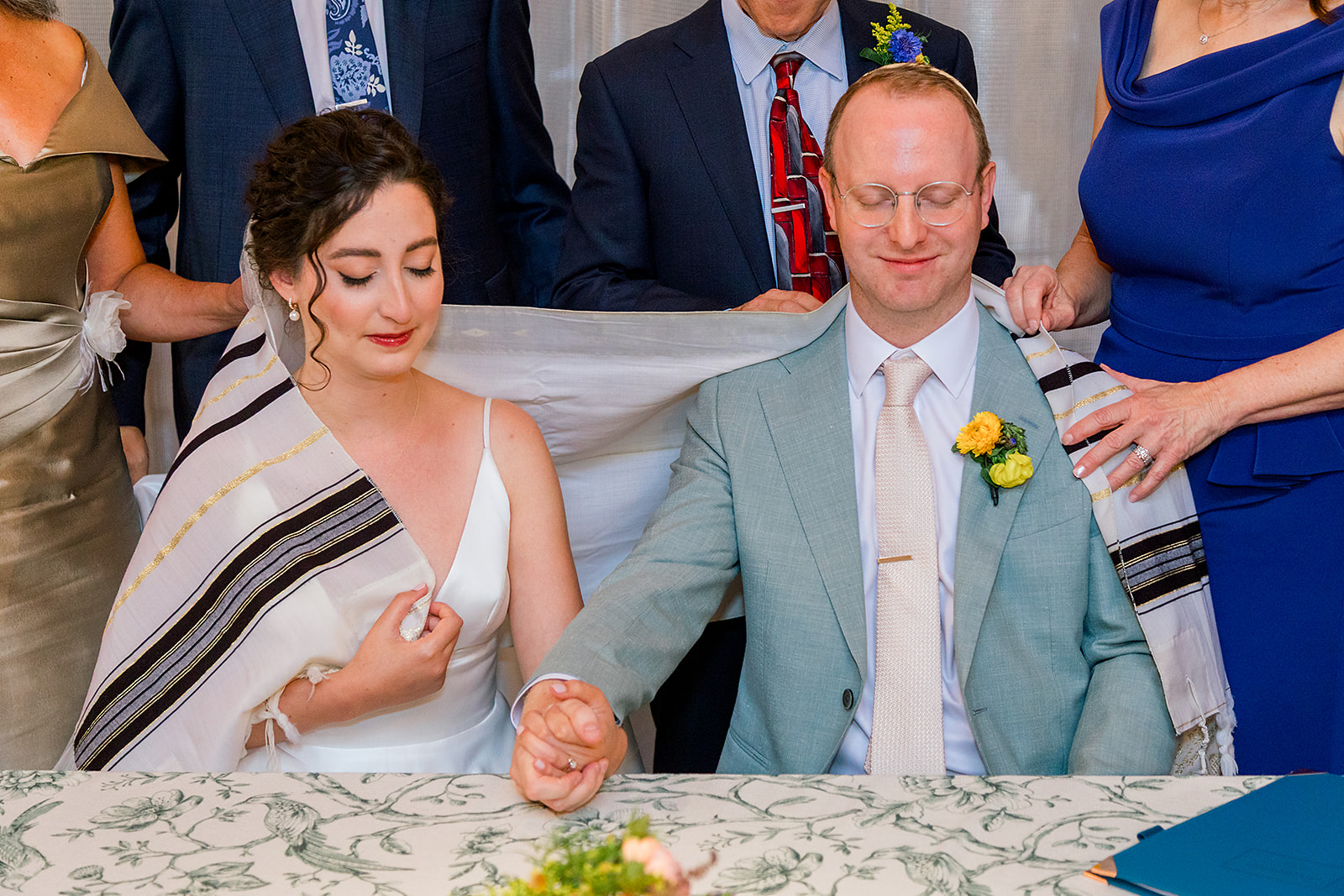 Newlyweds sit holding hands and praying with their parents hand on their backs during their Jewish wedding