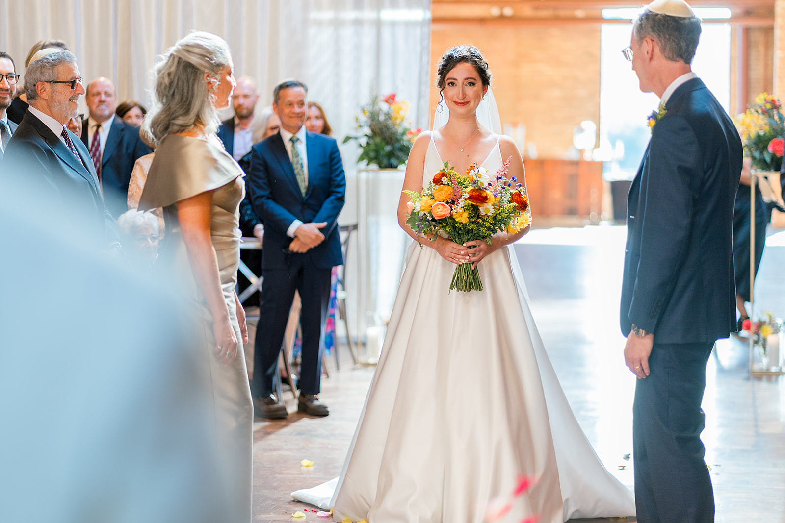 A bride smiles and holds back tears while walking down the aisle with mom and dad to her Jewish wedding ceremony