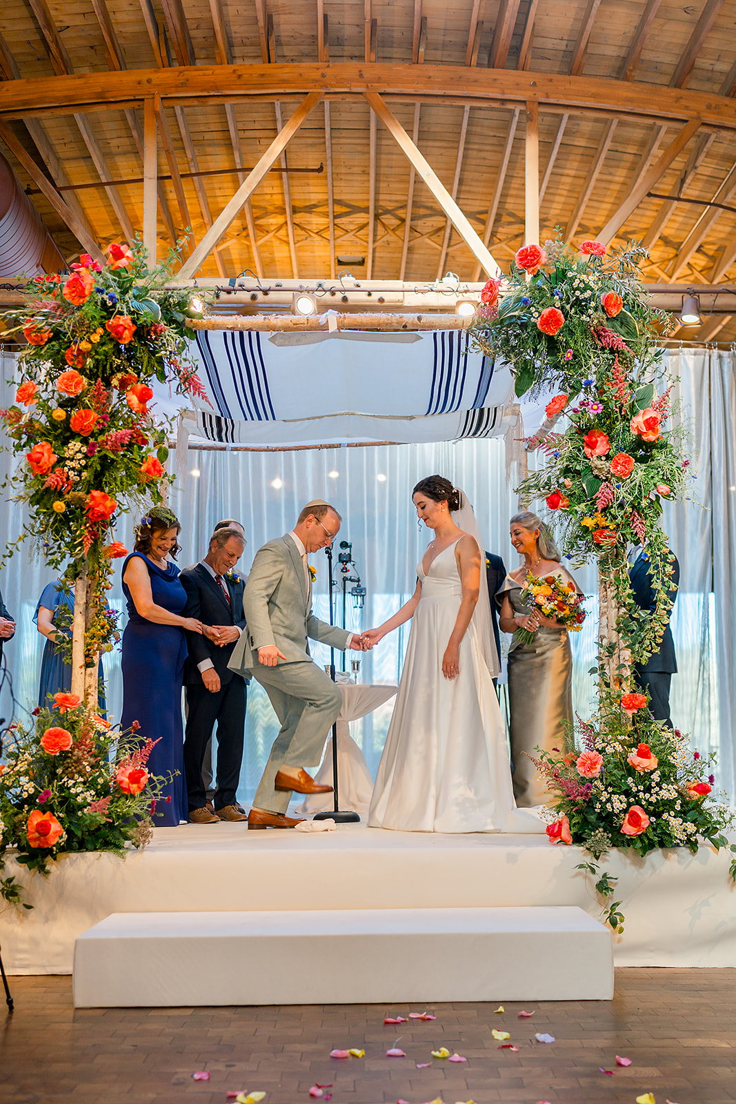 A groom in a green suit crushes the glass at the altar during his Jewish wedding while holding hands with the bride