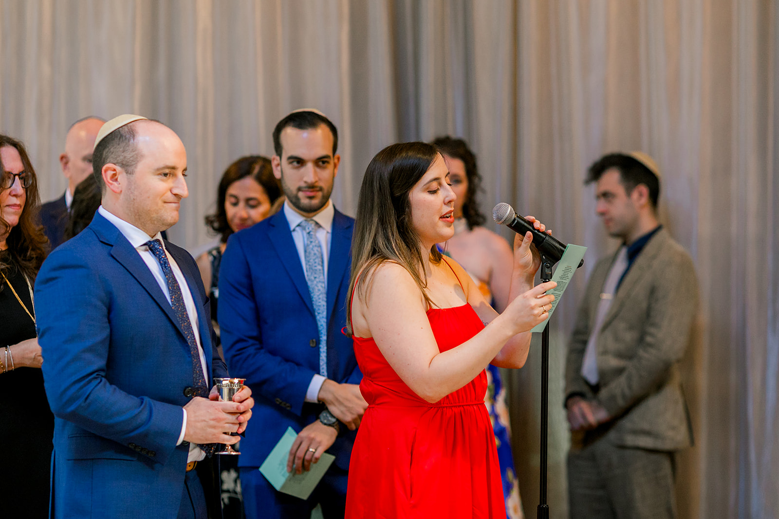 A woman in a red dress gives a speech at the microphone as others wait for their turn behind her
